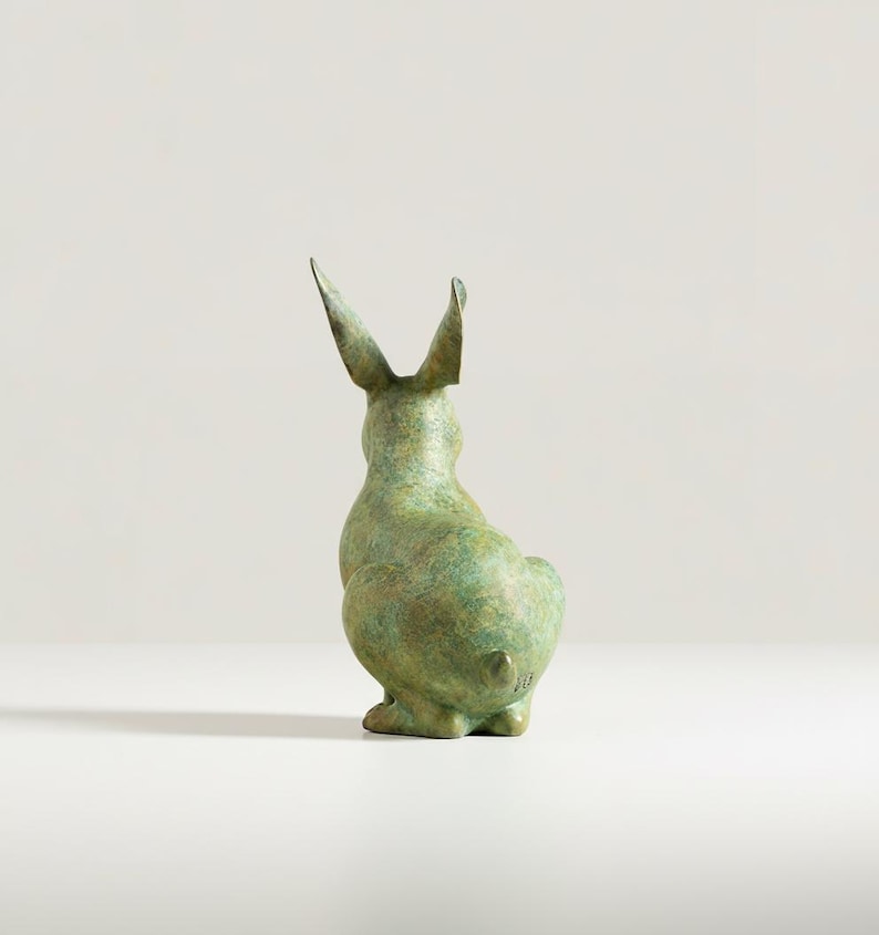 May include: A bronze-colored rabbit sculpture, viewed from the back, with long, upright ears. The sculpture has a textured surface and a slight green patina, suggesting age or weathering. The rabbit is in a seated position.