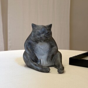 May include: A dark gray, seated cat figurine with a rounded, plump body and a smooth, matte finish. The cat is depicted in a relaxed, upright sitting position with its front paws resting near its belly and its eyes closed in a calm expression.