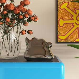 May include: A metallic, silver-colored decorative object with a rounded shape and a smooth, reflective surface. The object sits on a blue surface, with a vase of orange roses and a framed artwork in the background.
