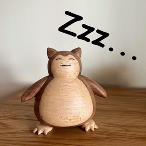 Puede incluir: Escultura de madera de un personaje de dibujos animados durmiendo, en tonos marrones y beige. La figura tiene los ojos cerrados y una pequeña sonrisa. Encima, las palabras "Zzz..." en negro.