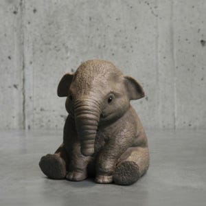 Puede incluir: Una pequeña figura de elefante marrón en posición sentada. El elefante tiene orejas grandes, una trompa y una superficie texturizada. La figura está hecha de un material similar a la piedra y está sobre un fondo gris.