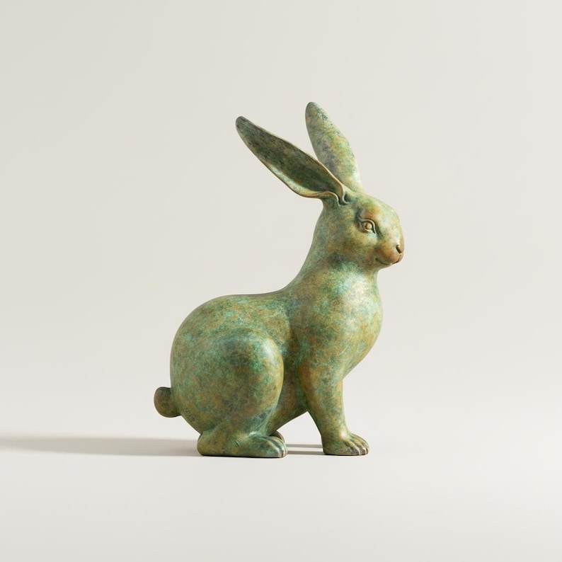 May include: A bronze-colored rabbit sculpture with a green patina. The rabbit is in a seated position with its ears erect. The sculpture has a textured surface and is set against a neutral background.