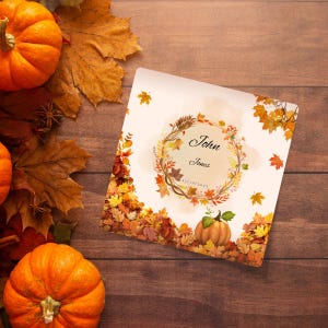 Op de afbeelding: Een vierkante uitnodigingskaart met een herfstthema, met een krans en een kleine pompoenillustratie. De kaart heeft de namen "John Jones" en de datum "11/10/2023". Oranje pompoenen en herfstbladeren omringen de kaart op een houten oppervlak.