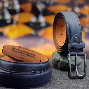 Op de afbeelding: Een donkerblauwe leren riem met een zilveren gesp. De riem heeft een gepersonaliseerde boodschap gegraveerd aan de binnenkant: "Happy third anniversary, Jonathan! Think of me when you take this off Love, Amy"