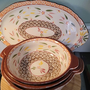 May include: A set of ceramic serving dishes with a floral design. The set includes a large oval platter and several bowls. The dishes have a white background with brown, yellow, pink, and green floral patterns and brown rims.