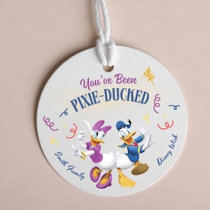 May include: A white round tag with a colourful design featuring Donald Duck and Daisy Duck. The tag says "You've Been Pixie-Ducked" with confetti and stars around the characters. The text "Smith Family" and "Disney Wish" are on the bottom of the tag.