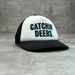 May include: A black and white trucker hat featuring a white front panel with the black text CATCHIN DEERS printed in a bold sans-serif font. The cap includes a black brim and black mesh side and back panels with a button on the top crown.