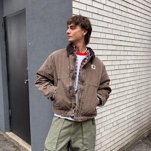 May include: A brown canvas work jacket with a dark brown corduroy collar and a small square logo patch on the chest, worn over a red and white plaid button-down shirt, a white t-shirt, and loose-fitting olive green cargo pants with large pockets.