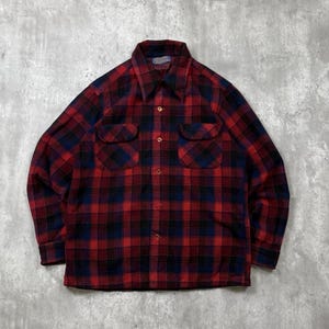 May include: A long-sleeve button-down shirt featuring a red and navy blue plaid pattern. The shirt includes a pointed collar, two chest pockets with buttoned flaps, and a front button closure. The fabric has a textured appearance throughout.