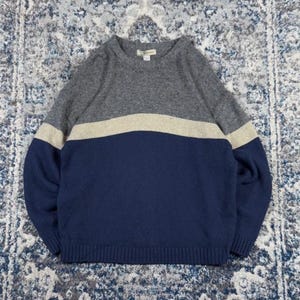 May include: A long-sleeved crew neck jumper featuring a colour-blocked design. The top section is charcoal grey, the middle is a horizontal beige stripe, and the bottom section is navy blue. The jumper has ribbed cuffs and a ribbed hemline.