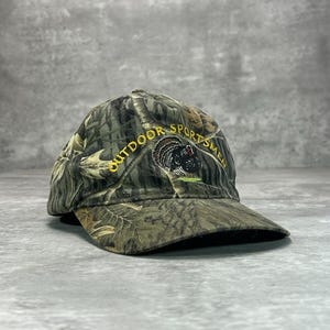 May include: Camouflage baseball cap with the text "OUTDOOR SPORTSMEN" in yellow lettering. The cap features a turkey graphic. The hat is made of a camouflage pattern fabric.
