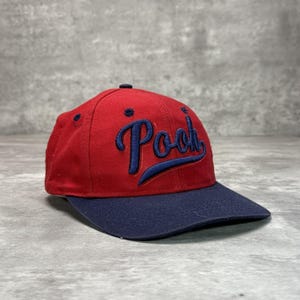 May include: A red baseball cap with a dark blue brim and a dark blue embroidered script logo that reads Pooh on the front. The cap features a structured crown with a dark blue button at the top and matching dark blue eyelets for ventilation.