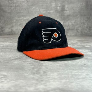 Op de afbeelding: Een zwarte honkbalpet met een geborduurd Philadelphia Flyers-logo op de voorzijde. De pet heeft een feloranje klep, een bijpassende oranje knoop op de kroon en oranje stiksels bij de oogjes. Het logo is wit met een oranje cirkel in het midden.