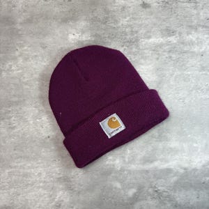 May include: A deep purple beanie hat with a folded brim. The hat features a small, rectangular patch with the Carhartt logo in white and gold. The hat is made of a knitted material and is suitable for cold weather.