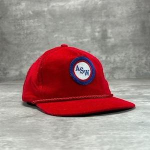 Puede incluir: Gorra de béisbol roja de pana con visera curva. La gorra presenta un parche circular con las letras "ASW" en blanco, rodeado por un borde azul y blanco. Un detalle de cuerda adorna el borde.