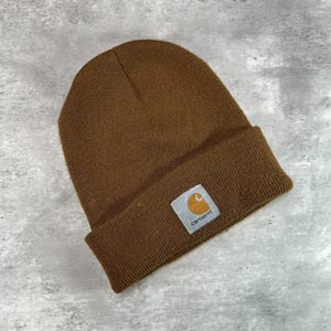 May include: A brown knit beanie with a folded cuff. A square white patch is sewn onto the center of the cuff, featuring the Carhartt brand name in black text and a yellow and orange circular logo graphic above the lettering.