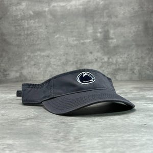 May include: A dark gray visor with a curved brim and a small, adjustable strap. The visor features a white and blue emblem on the front. The visor is designed to shield the eyes from the sun.