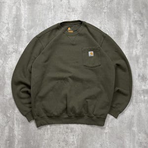 Op de afbeelding: Een olijfgroene sweater met ronde hals, lange mouwen, een geribbelde kraag en een borstzak aan de linkerkant. Een klein rechthoekig Carhartt-merklabel is op de zak genaaid. Het kledingstuk heeft een ontwerp met een relaxte pasvorm.