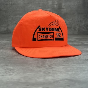 Op de afbeelding: Fel oranje baseballpet met een gebogen klep. De pet heeft zwarte tekst: "SKYDOME CHAMPION 92" met een honkbalafbeelding. De pet is gemaakt van lichtgewicht materiaal.