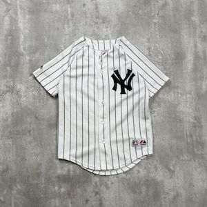 May include: White baseball jersey with black pinstripes and the New York Yankees logo on the chest. The jersey has short sleeves and a button-up front. A small logo is visible on the lower right.