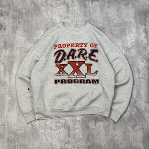 Può includere: Una felpa grigio chiaro con la scritta "PROPERTY OF DARE XXL PROGRAM" in lettere rosse e nere. La felpa ha un girocollo e maniche lunghe. Il tessuto sembra essere un materiale morbido e confortevole.