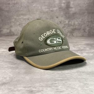 May include: An olive green baseball cap with a tan brim edge. The front features embroidered text reading GEORGE STRAIT, a central oval logo with the letters GS, and the text COUNTRY MUSIC FESTIVAL, accompanied by two small star icons.