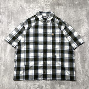 May include: A short-sleeve button-down shirt featuring a plaid pattern in shades of dark olive green, white, and black. The shirt includes a pointed collar, a front button placket, and a single chest pocket with a small Carhartt brand logo tag.