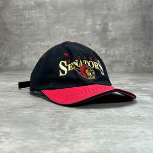 May include: A black baseball cap with a red brim and a red button on top. The front features the embroidered text Ottawa Senators in gold and white, accompanied by a red, gold, and black logo of a Roman centurion profile facing to the right.