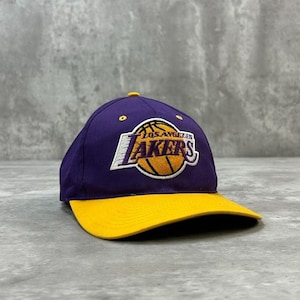 Peut inclure: Une casquette de baseball violette avec une visière jaune arborant le logo de l'équipe de basket-ball Los Angeles Lakers brodé sur le devant. Le logo comprend un ballon de basket jaune avec le texte Los Angeles Lakers en lettres violettes et blanches.