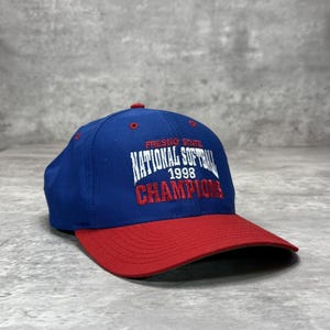 Op de afbeelding: Een blauwe honkbalpet met een rode klep, voorzien van wit en rood borduurwerk met de tekst FRESNO STATE NATIONAL SOFTBALL 1998 CHAMPIONS. De pet heeft een rode knoop aan de bovenkant en rode oogjes op de zijpanelen van de kroon.