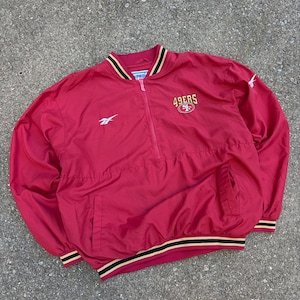 May include: A red long-sleeve pullover jacket featuring a half-zip closure, a ribbed collar with black and gold stripes, and matching striped cuffs. The front displays the text 49ERS with a logo, a Reebok logo, and a Pro Line tag at the neck.