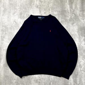 May include: A navy blue long-sleeve sweater featuring a crew neckline and a small red embroidered logo on the left chest. The garment includes a dark blue interior neck label with the text Polo Ralph Lauren and size information clearly visible.