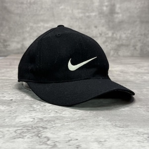 Op de afbeelding: Een zwarte honkbalpet met een wit geborduurd Nike swoosh-logo op het voorpaneel. De pet heeft een gestructureerde kroon, een gebogen klep en een knoopje aan de bovenkant. Het ontwerp is eenvoudig, met een effen zwarte stoffen constructie.
