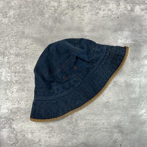 May include: A dark blue denim bucket hat with a brown brim. The hat has a classic design with a rounded crown and a downward-sloping brim. Two small metal rivets are visible on the hat's side.