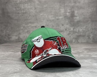 May include: Green baseball cap featuring a Coca-Cola polar bear wearing sunglasses and a racing suit. The cap has a black brim and the number "18" on the side. The Interstate Batteries logo is also visible.