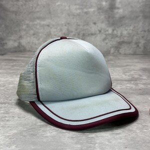 May include: A light gray trucker hat featuring a structured front panel and a breathable mesh back. The cap is accented with a dark maroon trim along the edges of the brim and the front panel, providing a distinct contrast to the light gray fabric.