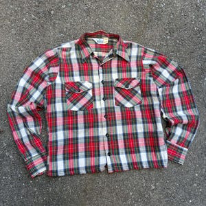 May include: A long-sleeve button-down shirt featuring a red, white, green, and black plaid pattern. The shirt includes a pointed collar, two chest pockets with buttoned flaps, and a front button closure. A Woolrich brand label is visible at the neck.