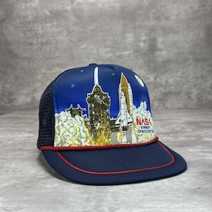 Può includere: Cappellino da camionista blu navy con retro in rete e visiera curva. Il pannello frontale presenta una grafica di un lancio dello space shuttle con la scritta "NASA Kennedy Space Center". Il cappello ha un dettaglio di corda rossa lungo il bordo.