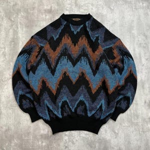 Op de afbeelding: Een patroon trui in zwart, blauw en bruin met een ronde hals. Het zigzagontwerp heeft tinten zwart, blauw en bruin. De manchetten en zoom zijn effen zwart.