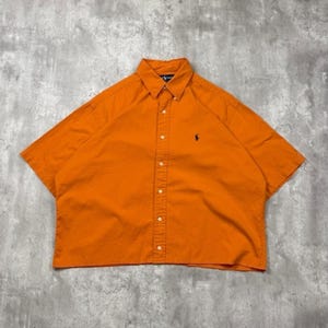 May include: A short-sleeved button-down shirt in a vibrant orange color. The shirt features a classic collar, a front button placket, and a small dark-colored embroidered logo of a person on a horse on the left side of the chest area.