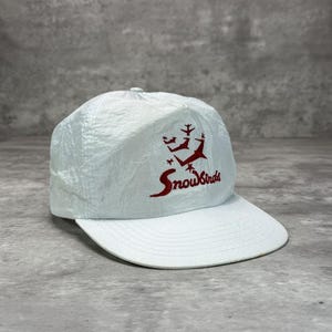 May include: A pale blue baseball cap with a white peak. The cap has the red text "Snowbirds" and a graphic of skiers and airplanes. The cap is made of a lightweight material, possibly nylon.