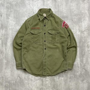 May include: An olive green button-down shirt with two chest pockets and a collar. It has a red patch on the right sleeve with the number 50 and text. There is a small text detail above the left pocket.