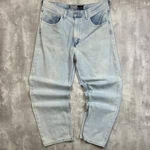 May include: Light blue denim jeans featuring a loose fit, a classic five-pocket design with metal rivets, a button and zipper fly closure, and a branded leather patch on the back waistband. The fabric has a faded wash with subtle whiskering.