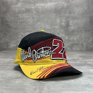 Può includere: Cappellino da baseball nero, giallo e rosso con il numero 2 e il nome "Ward Burton". Il cappellino ha una visiera curva e un bottone giallo. Il design include una firma sulla visiera.