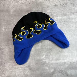 Op de afbeelding: Een blauwe en zwarte fleece muts met gele vlamaccenten. De muts heeft oorflappen en een gebogen ontwerp. De bovenkant is zwart, de onderkant en oorflappen zijn blauw. De vlammen zijn geel omlijnd.