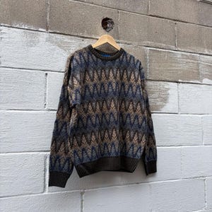Può includere: Un maglione a maniche lunghe con girocollo caratterizzato da un motivo geometrico a zigzag ripetuto nei toni del marrone scuro, beige e blu navy. Il capo presenta bordi in maglia a coste marrone scuro su colletto, polsini e orlo inferiore.