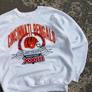 May include: A white long-sleeve sweatshirt featuring a graphic print that reads Cincinnati Bengals in orange letters. The design includes a football helmet, a red laurel wreath, and a banner stating 1988 AFC Champions and Super Bowl XXIII.