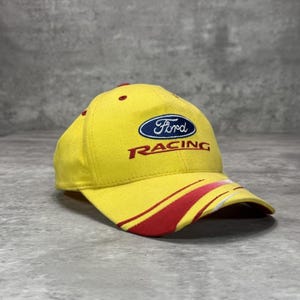 Peut inclure: Une casquette de baseball jaune vif arborant le logo Ford bleu et le mot RACING brodé en rouge sur le devant. La visière incurvée présente un motif à rayures graphiques rouge et blanc. La casquette possède des œillets rouges sur la calotte.