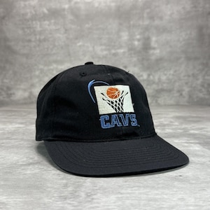 Op de afbeelding: Zwarte baseballcap met een basketbalringontwerp, een basketbal en het woord "CAVS" in blauw. De cap heeft een gebogen klep en een knoop aan de bovenkant. Het ontwerp staat op de voorkant.