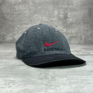 Op de afbeelding: Een grijze baseballpet met een zwarte klep. De pet heeft een rood Nike swoosh-logo en het woord "BASEBALL" in zwart geborduurd. De hoed is gemaakt van een textuurstof.